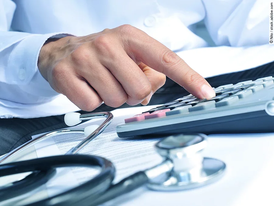 Die Hand eines Mannes in blauem Hemd tippt auf einem Taschenrechner. Daneben liegt ein Stethoskop.