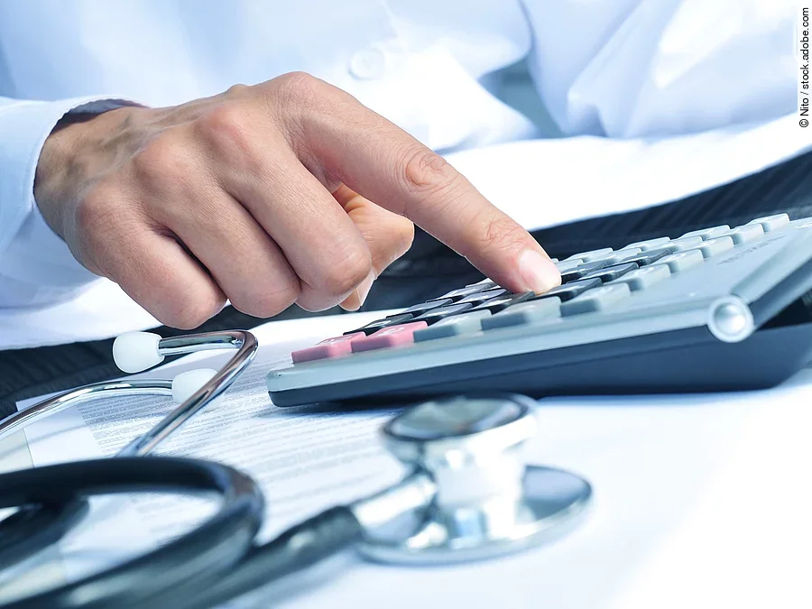 Die Hand eines Mannes in blauem Hemd tippt auf einem Taschenrechner. Daneben liegt ein Stethoskop.