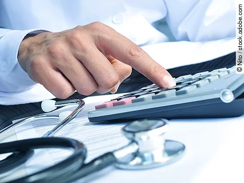 Die Hand eines Mannes in blauem Hemd tippt auf einem Taschenrechner. Daneben liegt ein Stethoskop.