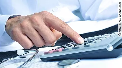 Taschenrechner Die Hand eines Mannes in blauem Hemd tippt auf einem Taschenrechner. Daneben liegt ein Stethoskop.