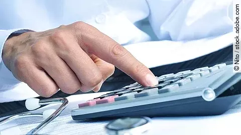 Die Hand eines Mannes in blauem Hemd tippt auf einem Taschenrechner. Daneben liegt ein Stethoskop.