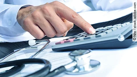 Die Hand eines Mannes in blauem Hemd tippt auf einem Taschenrechner. Daneben liegt ein Stethoskop.