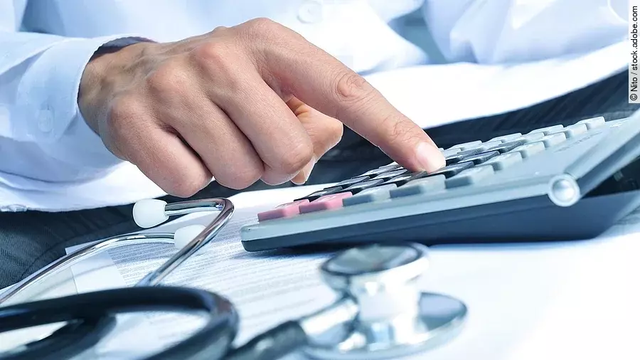 Die Hand eines Mannes in blauem Hemd tippt auf einem Taschenrechner. Daneben liegt ein Stethoskop.