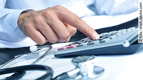 Die Hand eines Mannes in blauem Hemd tippt auf einem Taschenrechner. Daneben liegt ein Stethoskop.