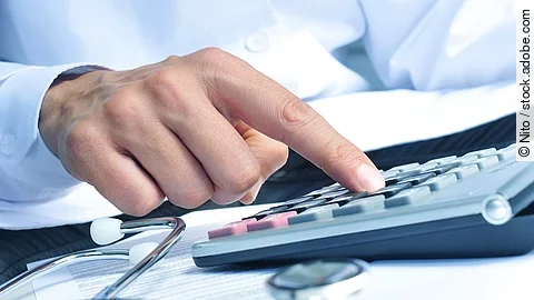 Die Hand eines Mannes in blauem Hemd tippt auf einem Taschenrechner. Daneben liegt ein Stethoskop.