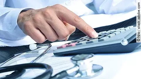 Die Hand eines Mannes in blauem Hemd tippt auf einem Taschenrechner. Daneben liegt ein Stethoskop.