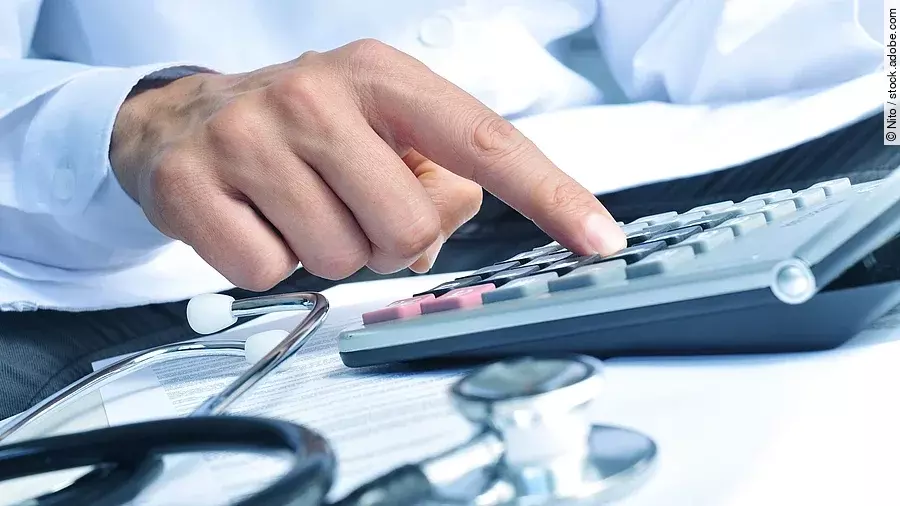Die Hand eines Mannes in blauem Hemd tippt auf einem Taschenrechner. Daneben liegt ein Stethoskop.