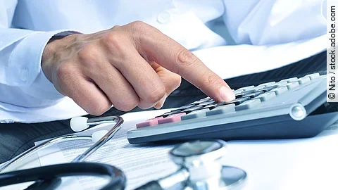 Die Hand eines Mannes in blauem Hemd tippt auf einem Taschenrechner. Daneben liegt ein Stethoskop.