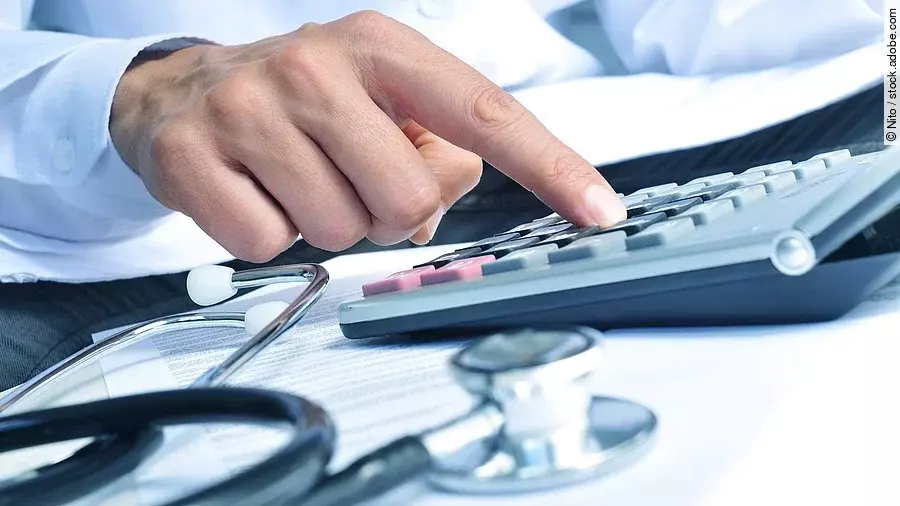 Die Hand eines Mannes in blauem Hemd tippt auf einem Taschenrechner. Daneben liegt ein Stethoskop.