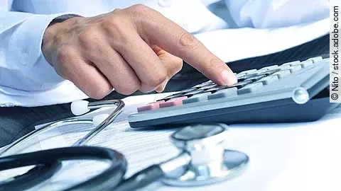 Die Hand eines Mannes in blauem Hemd tippt auf einem Taschenrechner. Daneben liegt ein Stethoskop.