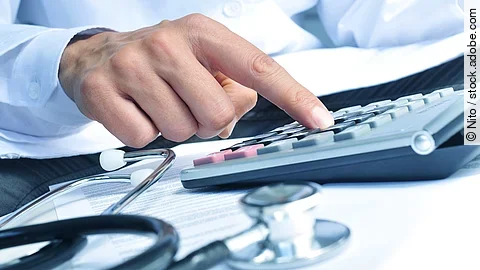 Die Hand eines Mannes in blauem Hemd tippt auf einem Taschenrechner. Daneben liegt ein Stethoskop.