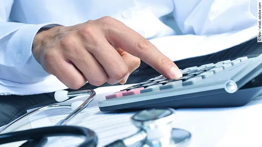 Die Hand eines Mannes in blauem Hemd tippt auf einem Taschenrechner. Daneben liegt ein Stethoskop.
