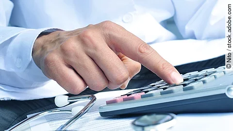 Die Hand eines Mannes in blauem Hemd tippt auf einem Taschenrechner. Daneben liegt ein Stethoskop.