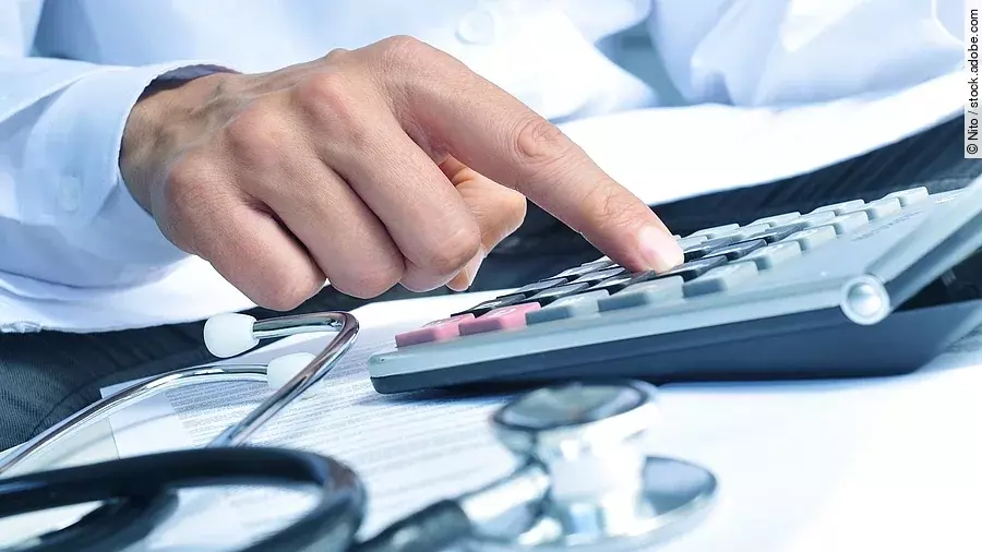 Die Hand eines Mannes in blauem Hemd tippt auf einem Taschenrechner. Daneben liegt ein Stethoskop.