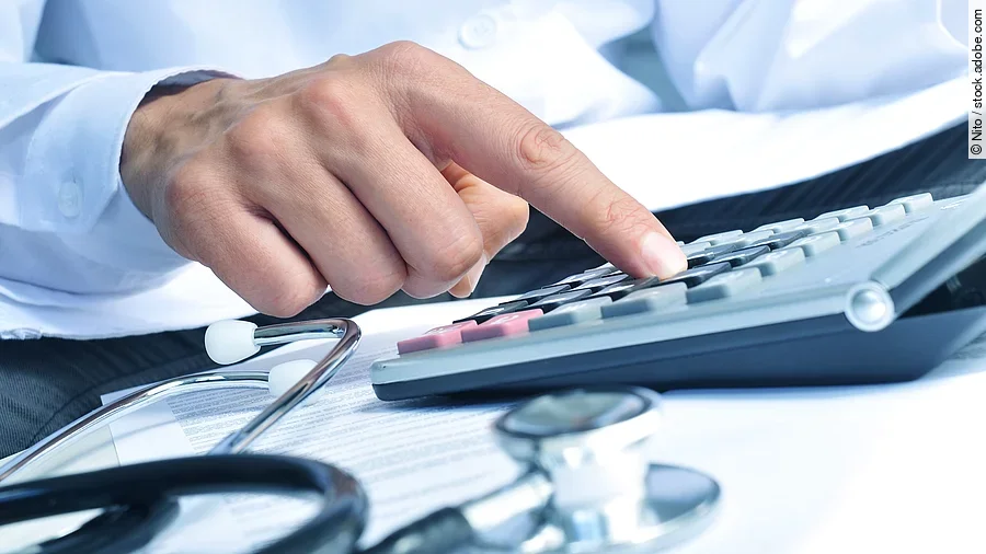 Die Hand eines Mannes in blauem Hemd tippt auf einem Taschenrechner. Daneben liegt ein Stethoskop.