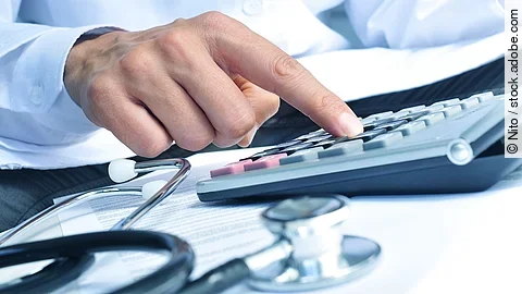 Taschenrechner Die Hand eines Mannes in blauem Hemd tippt auf einem Taschenrechner. Daneben liegt ein Stethoskop.