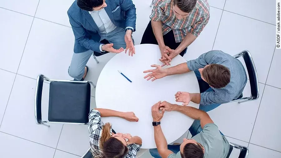Personen sitzen an einem kleinen runden Tisch und diskutieren. Ein Sitzplatz ist frei.