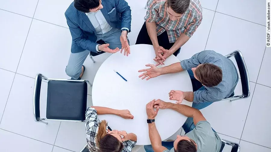 Personen sitzen an einem kleinen runden Tisch und diskutieren. Ein Sitzplatz ist frei.