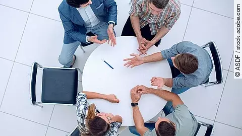 Personen sitzen an einem kleinen runden Tisch und diskutieren. Ein Sitzplatz ist frei.
