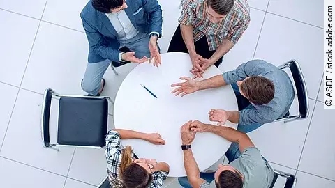 Personen sitzen an einem kleinen runden Tisch und diskutieren. Ein Sitzplatz ist frei.