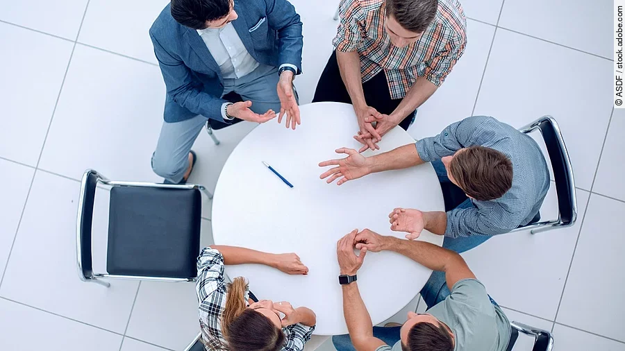 Personen sitzen an einem kleinen runden Tisch und diskutieren. Ein Sitzplatz ist frei.