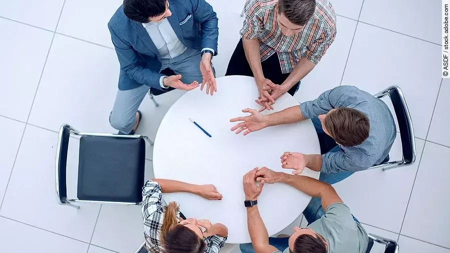 Personen sitzen an einem kleinen runden Tisch und diskutieren. Ein Sitzplatz ist frei.