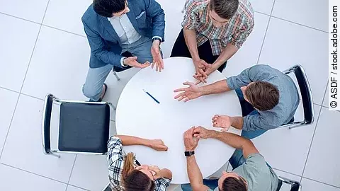 Personen sitzen an einem kleinen runden Tisch und diskutieren. Ein Sitzplatz ist frei.