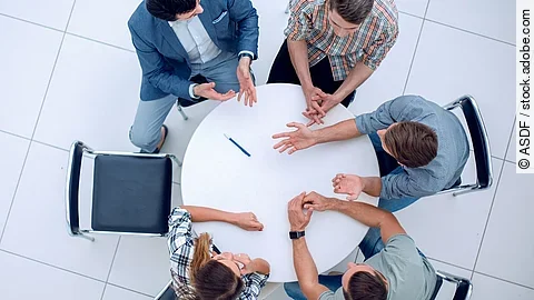 Personen sitzen an einem kleinen runden Tisch und diskutieren. Ein Sitzplatz ist frei.