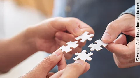 Vier Hände halten jeweils ein weißes Puzzleteil und setzen sie zusammen.