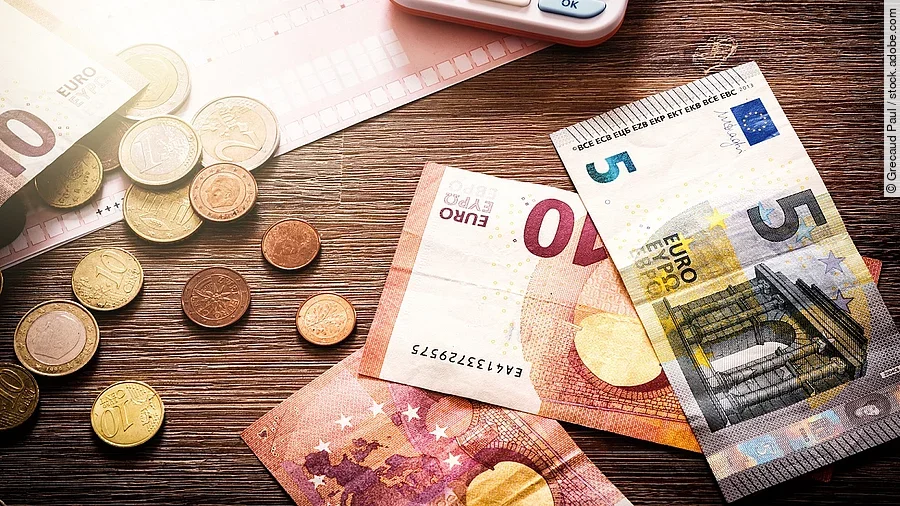 Mehrere Geldmünzen, Banknoten und ein Taschenrechner liegen verteilt auf dem Tisch.