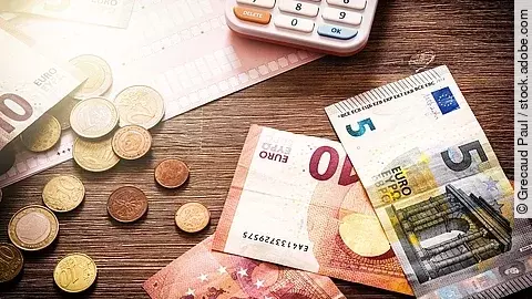 Mehrere Geldmünzen, Banknoten und ein Taschenrechner liegen verteilt auf dem Tisch.