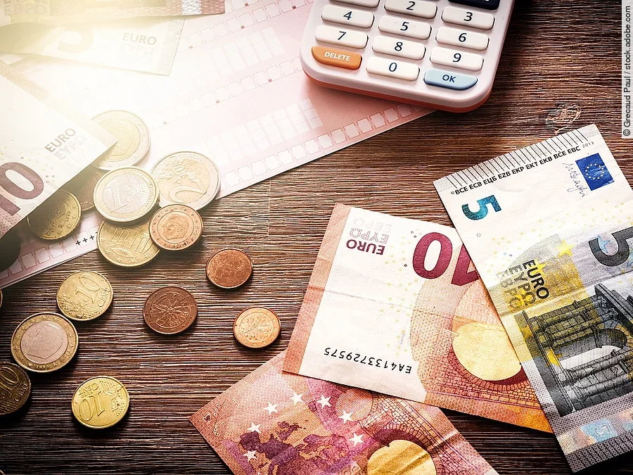Mehrere Geldmünzen, Banknoten und ein Taschenrechner liegen verteilt auf dem Tisch.