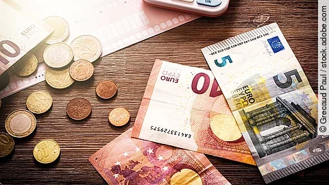 Mehrere Geldmünzen, Banknoten und ein Taschenrechner liegen verteilt auf dem Tisch.