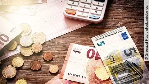 Mehrere Geldmünzen, Banknoten und ein Taschenrechner liegen verteilt auf dem Tisch.