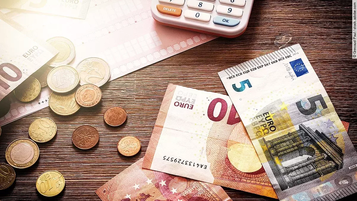 Mehrere Geldmünzen, Banknoten und ein Taschenrechner liegen verteilt auf dem Tisch.