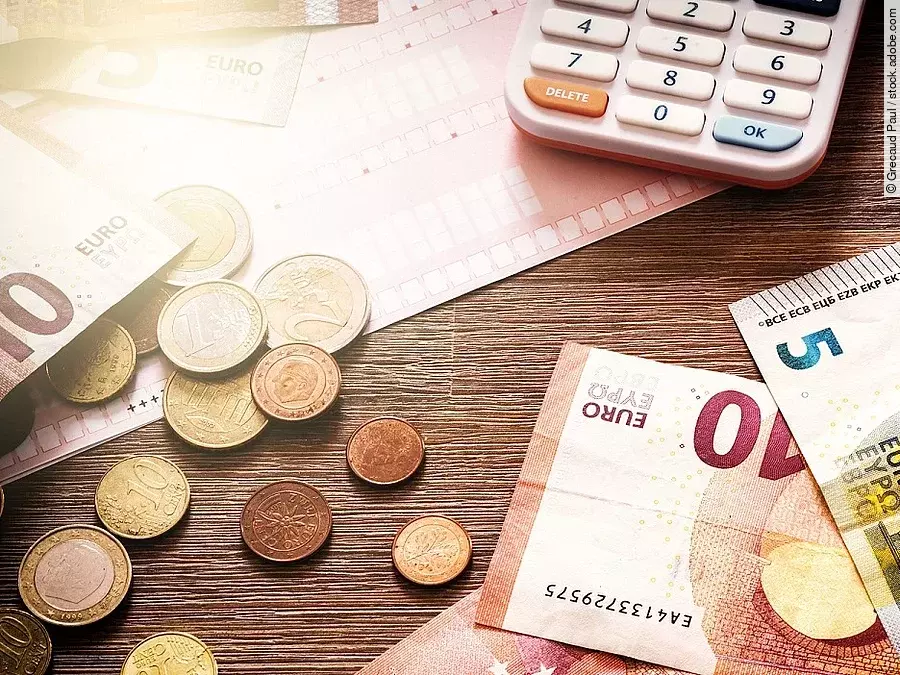 Mehrere Geldmünzen, Banknoten und ein Taschenrechner liegen verteilt auf dem Tisch.