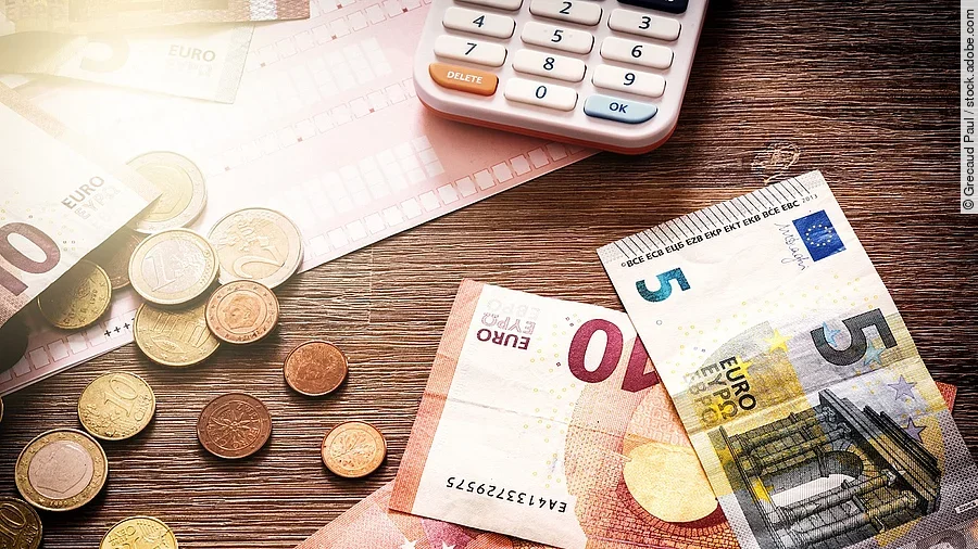 Mehrere Geldmünzen, Banknoten und ein Taschenrechner liegen verteilt auf dem Tisch.