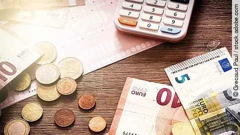 Mehrere Geldmünzen, Banknoten und ein Taschenrechner liegen verteilt auf dem Tisch.