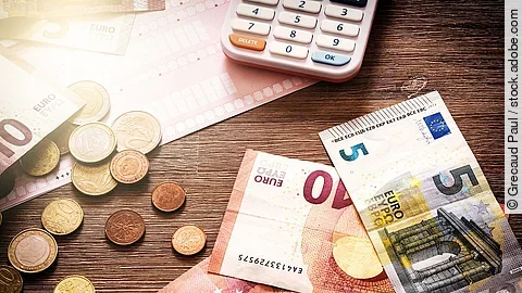 Mehrere Geldmünzen, Banknoten und ein Taschenrechner liegen verteilt auf dem Tisch.
