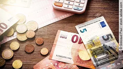 Mehrere Geldmünzen, Banknoten und ein Taschenrechner liegen verteilt auf dem Tisch.