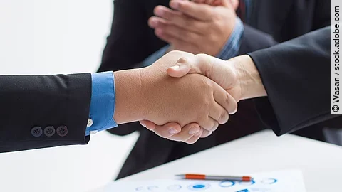 Zwei Geschäftsleute verüben einen Handschlag. Im Hintergrund applaudiert eine Person.