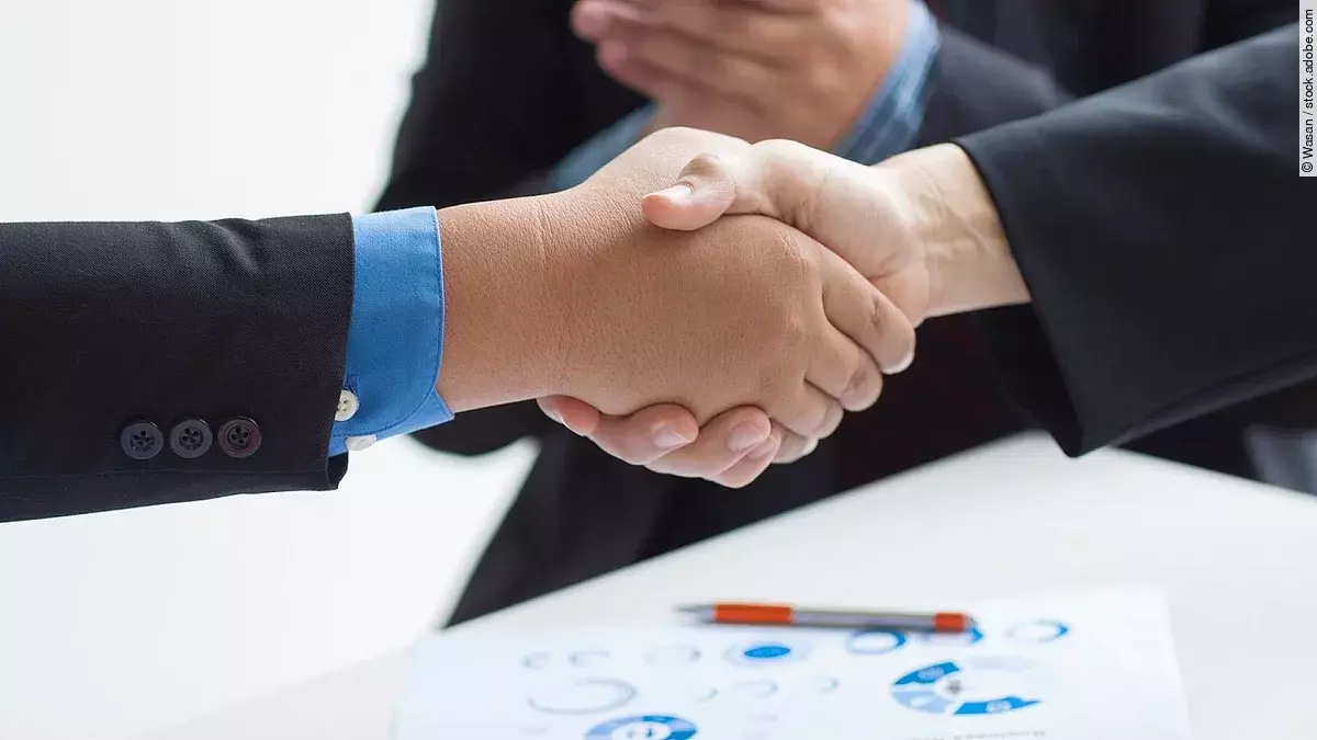 Zwei Geschäftsleute verüben einen Handschlag. Im Hintergrund applaudiert eine Person.