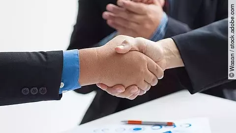 Zwei Geschäftsleute verüben einen Handschlag. Im Hintergrund applaudiert eine Person.