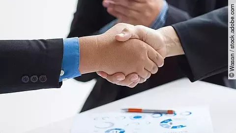 Zwei Geschäftsleute verüben einen Handschlag. Im Hintergrund applaudiert eine Person.