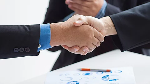 Zwei Geschäftsleute verüben einen Handschlag. Im Hintergrund applaudiert eine Person.