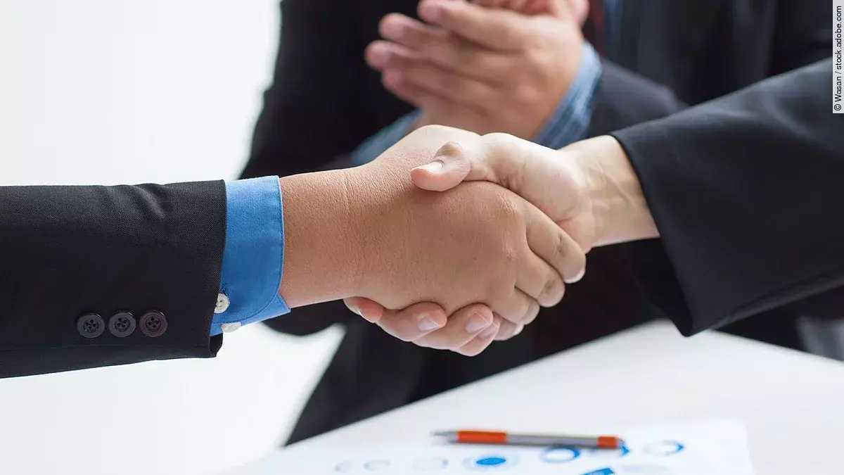 Zwei Geschäftsleute verüben einen Handschlag. Im Hintergrund applaudiert eine Person.