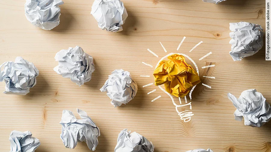 Viele zusammengeknüllte Blätter Papier. Eines davon in gelber Farbe und umrahmt wie eine leuchtende Glühbirne.