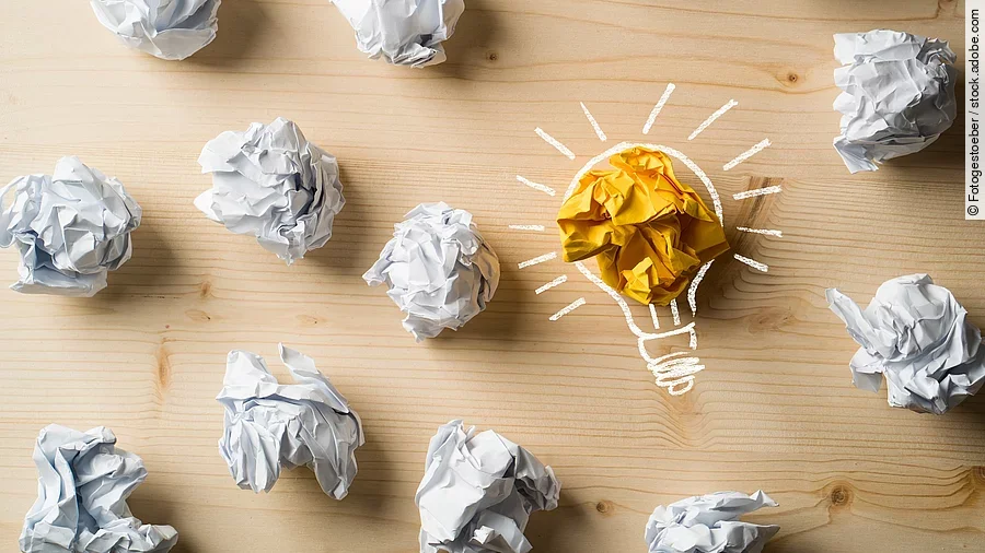 Viele zusammengeknüllte Blätter Papier. Eines davon in gelber Farbe und umrahmt wie eine leuchtende Glühbirne.