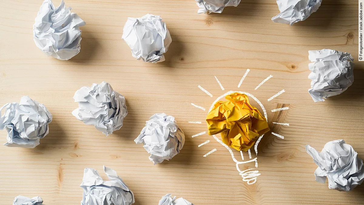 Viele zusammengeknüllte Blätter Papier. Eines davon in gelber Farbe und umrahmt wie eine leuchtende Glühbirne.