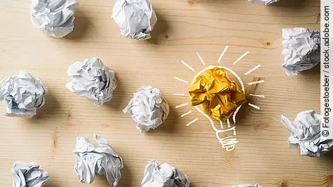 Viele zusammengeknüllte Blätter Papier. Eines davon in gelber Farbe und umrahmt wie eine leuchtende Glühbirne.